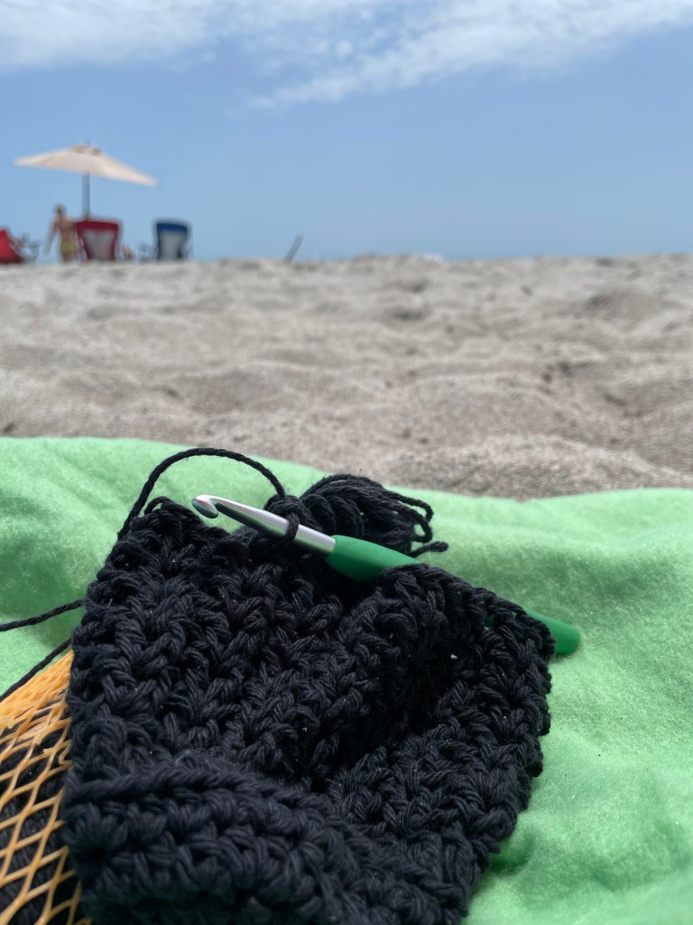 An image of a bottle holder in process on a beach. You can see a green blanket, a black bit of yarn and the horizon of sand and blue sky.