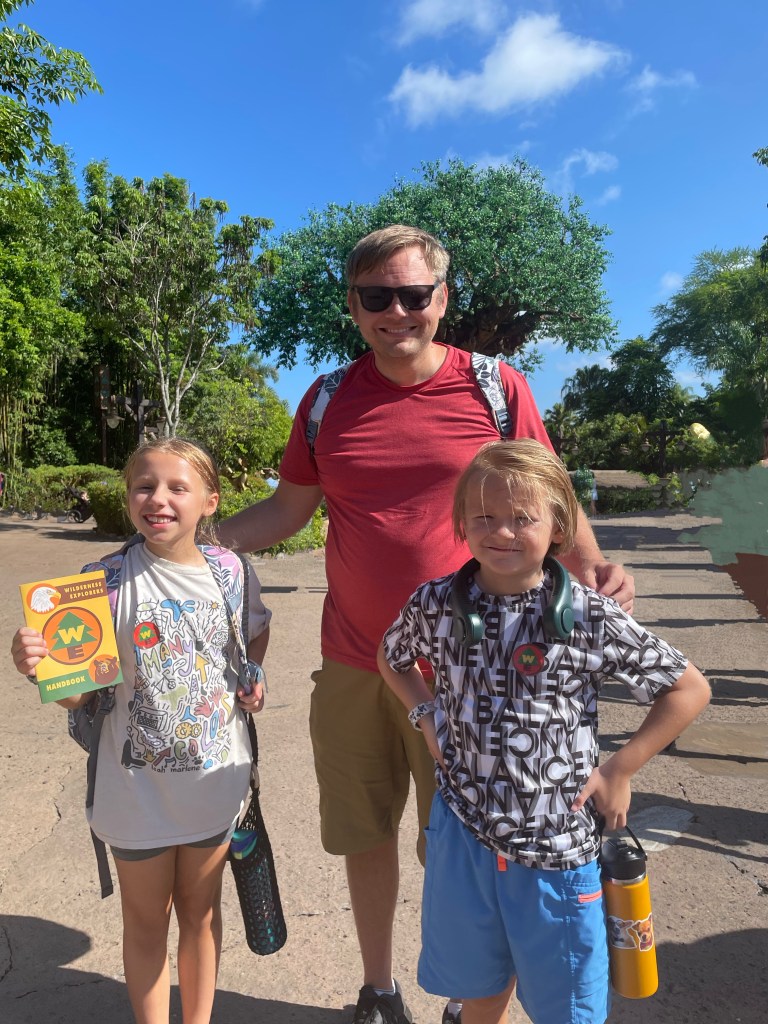 A family poses at Animal Kingdom at Walt Disney World. There is a man and two small children, one female, one male. The female child is holding a water bottle in a yarn case.